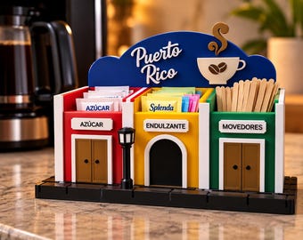 Puede incluir: Un organizador de estación de café con varios compartimentos, una parte superior azul con el texto Puerto Rico y un icono de taza. Tiene tres secciones etiquetadas como Azúcar, Endulzante y Movedores, diseñadas como edificios coloridos.