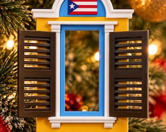 May include: A decorative hanging ornament shaped like a yellow window frame with open dark brown shutters. The top features a blue arch with a small Puerto Rican flag. The ornament is suspended by a gold ribbon and has a white frame border.