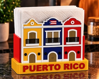 Puede incluir: Un servilletero con la fachada colorida de tres edificios en amarillo, azul y rojo. El edificio central muestra una pequeña bandera de Puerto Rico. La base es amarilla con el texto PUERTO RICO en letras rojas. Contiene servilletas blancas.