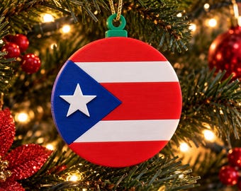 Puede incluir: Un adorno navideño redondo y plano con la bandera de Puerto Rico. El diseño incluye franjas horizontales rojas y blancas, un triángulo azul en el lado izquierdo y una estrella blanca centrada dentro del triángulo azul. Tiene una argolla verde.