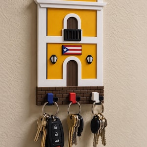 May include: A wall-mounted key holder shaped like a yellow colonial building with a white frame, a balcony, and a Puerto Rican flag. The base features a brown brick pattern with three hooks in blue, red, and white, each holding a set of metal keys.