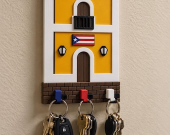 Puede incluir: Un llavero de pared con forma de edificio colonial amarillo con marco blanco, balcón y la bandera de Puerto Rico. La base presenta un patrón de ladrillos marrones con tres ganchos en azul, rojo y blanco, cada uno con un juego de llaves.