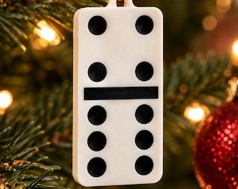 Puede incluir: Un adorno de dominó blanco con puntos negros y una barra central negra, que cuenta con un pequeño lazo en la parte superior para colgar. El dominó muestra un patrón de dos-seis y cuelga de un cordón dorado fino ante un árbol.