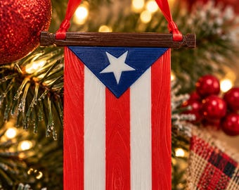 Puede incluir: Un adorno colgante con la forma de la bandera de Puerto Rico, que presenta un triángulo azul con una estrella blanca y franjas verticales alternas en rojo y blanco. La bandera cuelga de una barra horizontal marrón mediante una cinta roja.