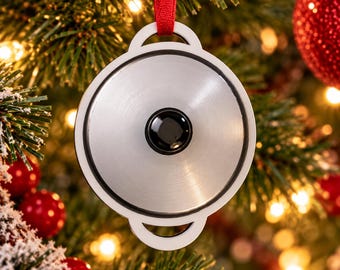 May include: A circular silver-toned ornament shaped like a cooking pot lid with a black central knob and a white outer frame. The ornament is suspended by a red ribbon and features a metallic finish with a thin black circular border.
