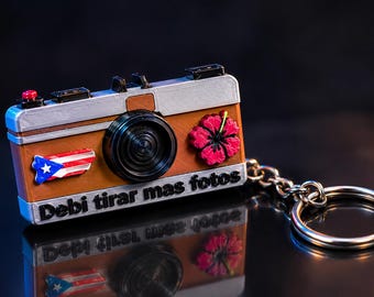 Puede incluir: Un llavero con forma de cámara marrón y plateada que presenta un objetivo negro, una flor de hibisco roja y un emblema de la bandera de Puerto Rico. El texto "Debi tirar mas fotos" está impreso en negro en el frente de la cámara.