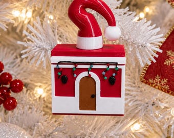 Puede incluir: Un adorno navideño cuadrado de color rojo y blanco con una puerta marrón, una guirnalda de luces multicolores y un gorro de Papá Noel rojo con un pompón blanco en la parte superior. El adorno cuelga de un cordón dorado en un árbol blanco.
