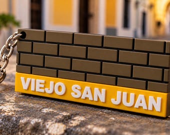 Puede incluir: Un llavero rectangular que presenta un patrón de pared de ladrillos de color verde oliva oscuro en la sección superior y una sección inferior de color amarillo brillante con el texto blanco en relieve VIEJO SAN JUAN.