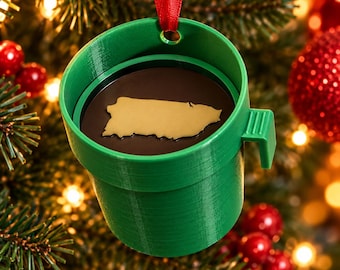 Puede incluir: Un adorno navideño verde con forma de taza cilíndrica que presenta una base interior de color marrón oscuro con un mapa dorado de Puerto Rico en el centro. El adorno tiene una pequeña asa lateral y una cinta roja para colgar.