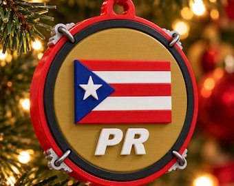 Puede incluir: Un adorno navideño redondo en forma de tambor con un centro dorado que muestra la bandera de Puerto Rico y las letras blancas PR. El adorno tiene un borde exterior rojo con herrajes plateados y una cinta roja.