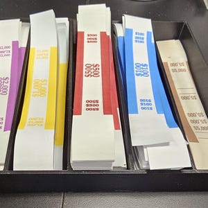 May include: A black tray filled with stacks of banded currency. The bands are color-coded, with denominations of $100, $500, $1,000, $2,000, and $5,000 visible. The bands are white with colored text.