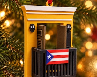 May include: A yellow architectural ornament featuring a balcony with a black railing, a Puerto Rican flag, two open brown doors, and two small black wall lamps. The piece is topped with a white cornice and hangs from a red ribbon loop.
