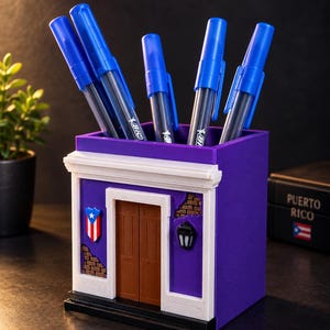 May include: A purple square pencil holder designed like a building facade with a brown door, white trim, a small Puerto Rican flag emblem, and a black lantern. Five blue Bic pens are inserted into the top opening of the container.