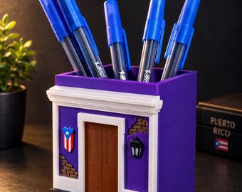 May include: A purple square pencil holder designed like a building facade with a brown door, white trim, a small Puerto Rican flag emblem, and a black lantern. Five blue Bic pens are inserted into the top opening of the container.