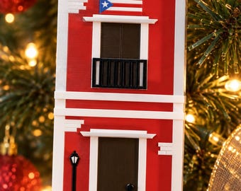 Puede incluir: Un adorno colgante rectangular rojo y blanco con forma de edificio de dos plantas, con puerta marrón oscuro, balcón negro, una pequeña farola negra y una bandera de Puerto Rico sobre la ventana superior, suspendido por una cinta roja.