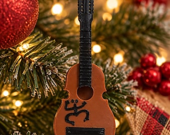 Puede incluir: Un adorno navideño con forma de guitarra marrón con mástil y clavijero negros. El cuerpo presenta un símbolo de rana Coquí negro y una barra rectangular negra horizontal. Una cinta roja está unida a la parte superior para colgar.