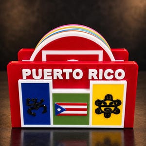 May include: A red coaster holder featuring the text PUERTO RICO in white letters. The front displays a blue square with a black coqui, a green square with a miniature Puerto Rican flag, and a yellow square with a black sun symbol. A stack of colorful round coasters is visible inside.