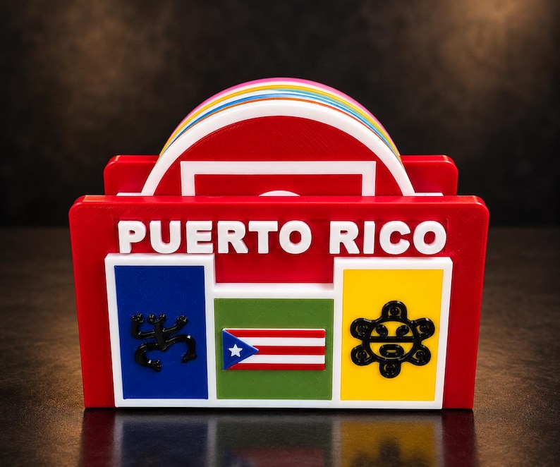 May include: A red coaster holder featuring the text PUERTO RICO in white letters. The front displays a blue square with a black coqui, a green square with a miniature Puerto Rican flag, and a yellow square with a black sun symbol. A stack of colorful round coasters is visible inside.