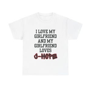 Puede incluir: Una camiseta blanca de manga corta con texto negro que dice I LOVE MY GIRLFRIEND AND MY GIRLFRIEND LOVES, seguido del nombre J-HOPE escrito en una fuente roja en negrita con un contorno negro centrado en la parte delantera de la prenda.