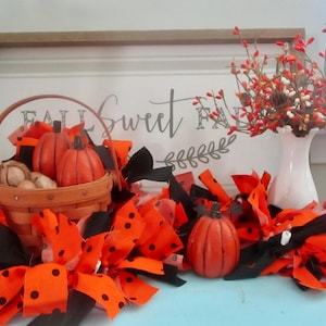 May include: A fall-themed display featuring a wooden basket with orange pumpkins and decorative gourds. The arrangement includes a garland of orange and black fabric with polka dots, a white vase with red berries, and a sign that says "Fall Sweet Fall."