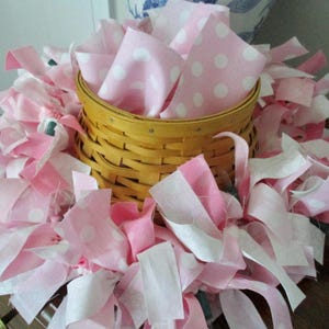 May include: A pink and white fabric wreath with a woven basket in the center. The fabric is cut into strips and tied together to create a fluffy wreath.