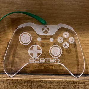 Pode incluir: Enfeite transparente de acrílico em forma de controle Xbox com detalhes gravados, incluindo botões, direcional e o texto "2022 BOSTON". Uma fita verde está presa para pendurar. Ideal para gamers.