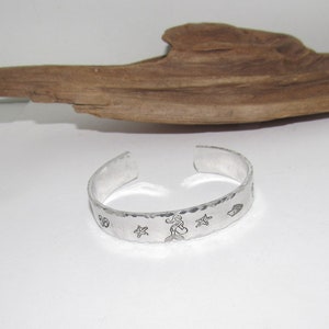 May include: Silver bracelet with a hammered finish and engraved mermaid, starfish, and seashell designs.