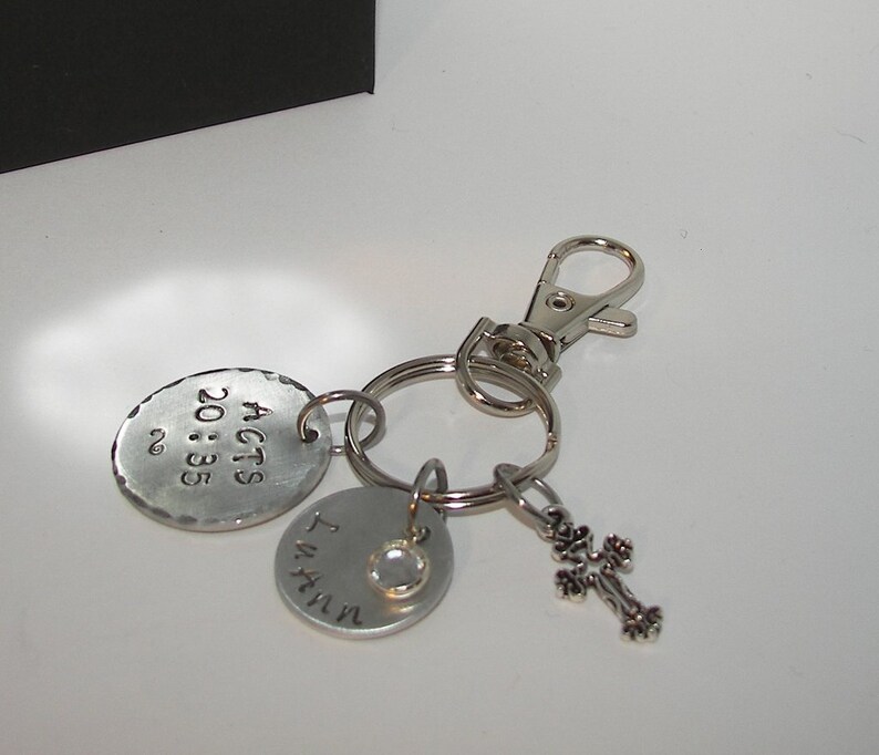K&ouml;nnte beinhalten: Silberner Schl&uuml;sselanh&auml;nger mit drei Anh&auml;ngern. Der gr&ouml;&szlig;te Anh&auml;nger ist rund und mit "ACTS 20:35" graviert. Der zweite Anh&auml;nger ist rund und mit "I WILL" graviert. Der dritte Anh&auml;nger ist ein kleines silbernes Kreuz.