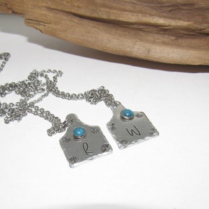May include: Two silver necklaces with turquoise stones and personalized tags. The tags are shaped like a cow tag and have the letters "R" and "W" engraved on them.