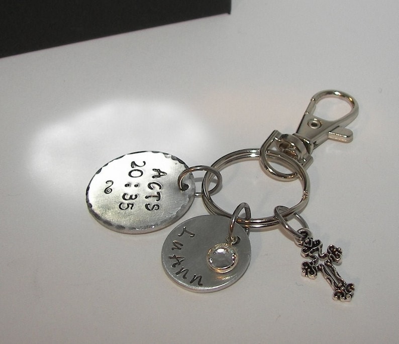 K&ouml;nnte beinhalten: Silberner Schl&uuml;sselanh&auml;nger mit drei Anh&auml;ngern. Der erste Anh&auml;nger ist ein rundes Metallschild mit dem Text "ACTS 20:35". Der zweite Anh&auml;nger ist ein rundes Metallschild mit dem Text "I AM IN". Der dritte Anh&auml;nger ist ein kleines silbernes Kreuz.