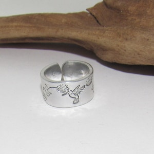 May include: A silver ring with an engraved design of a hummingbird flying near a flower.