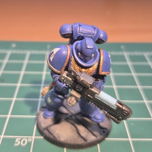 May include: A miniature blue and gold armoured figure holding a large gun. The figure has a helmet and shoulder pads with white and gold accents. The base is a stone-like texture. The figure is on a green cutting mat.