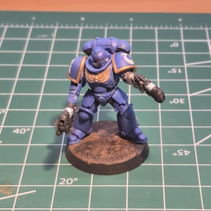 May include: A miniature blue and gold space marine figure, holding two guns. The figure stands on a brown circular base, against a green cutting mat background.