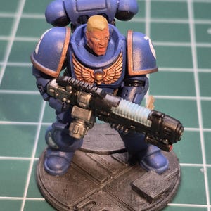 May include: A miniature figurine of a space marine in blue armour, holding a black and silver weapon. The figurine has a blonde head and is standing on a grey base. The armour features gold accents and white wing emblems.