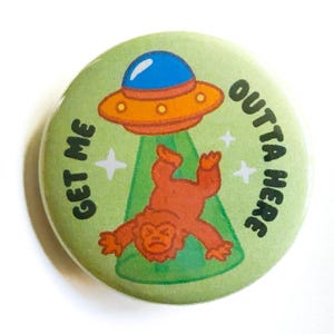 May include: A round, green button with a cartoon illustration of a UFO abducting a brown Bigfoot. The text "GET ME OUTTA HERE" is printed around the edge of the button. The UFO is orange and blue.