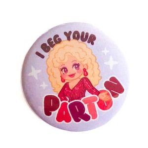 Op de afbeelding: Een ronde, lichtpaarse knoop met de tekst "I BEG YOUR PARDON" in rode en witte letters. De knoop toont een cartoonillustratie van een vrouw met blond krullend haar en een rode jurk.
