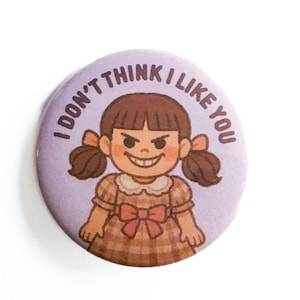 May include: A round, lavender button with the text "I DON'T THINK I LIKE YOU" encircling a cartoon girl with a stern expression. The girl has brown hair in pigtails and wears a plaid dress with a bow.