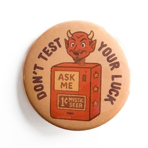 May include: A round, tan button with a cartoon devil and the text "DON'T TEST YOUR LUCK". The button features a red fortune-telling machine with the words "ASK ME" and "MYSTIC SEER".