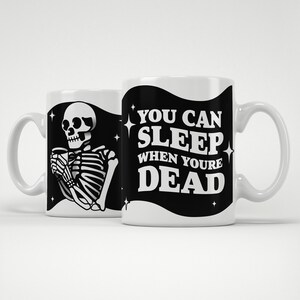 May include: Two white ceramic mugs with black and white graphics. One mug features a skeleton holding a cup, the other has the text "YOU CAN SLEEP WHEN YOU'RE DEAD". Both mugs have a curved handle.