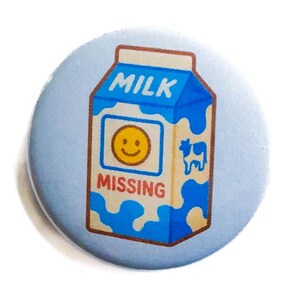 May include: A round, light blue button featuring a cartoon milk carton design. The carton is blue and white with the word "MILK" at the top and the word "MISSING" at the bottom. A smiling face is in the center.