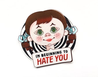 May include: A sticker featuring a cartoon girl with brown hair in pigtails, green eyes, and freckles. The girl is set against a black and white striped background. The text "I'M BEGINNING TO HATE YOU" is displayed on a white sign.