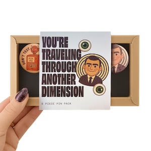 May include: A 5-piece pin pack in a tan box. The pins feature retro-style illustrations and text, including "YOU'RE TRAVELING THROUGH ANOTHER DIMENSION" and a cartoon man. One pin has a devil and the text "DON'T TEST".
