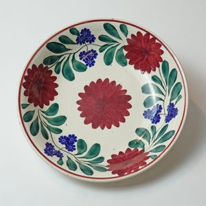 Può includere: Un piatto rotondo in ceramica color crema con bordo rosso e design floreale. Il piatto presenta grandi fiori rossi, foglie verdi e grappoli di fiori blu. Il fiore centrale è anch'esso rosso.