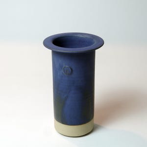 May include: A cylindrical ceramic vase in a deep blue hue, featuring a wide, flat rim and a lighter beige base. The vase has a subtle embossed design and a smooth, matte finish. The vase is approximately 20 cm tall.