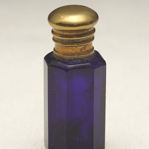 Puede incluir: Pequeña botella antigua de vidrio azul cobalto con cuerpo facetado y tapón de latón. La botella tiene forma hexagonal y se mantiene erguida. El tapón de latón es de diseño redondo y plano.