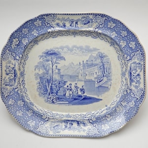 May include: An antique blue and white ceramic serving platter. The oval platter features a detailed landscape scene in the center, surrounded by a floral and vine border. The edges have a scalloped design.