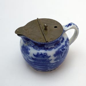 May include: A small, round, blue and white ceramic creamer with a metal lid. The creamer has a classic blue and white pattern, a handle and a spout. The metal lid is hinged and has a small knob.
