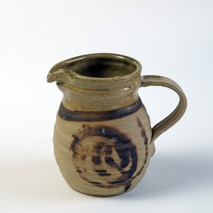 May include: A handmade ceramic pitcher with a spout and handle. The pitcher is tan with a dark brown band and a decorative swirl design. The rim has a green hue.