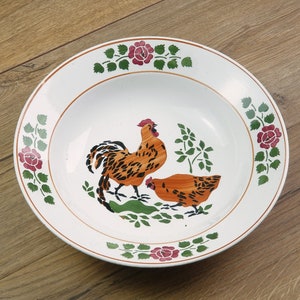 Op de afbeelding: Witte keramische serveerschaal met een haan- en kippenontwerp. De kom heeft een bruine rand en is versierd met een haan en een kip in tinten oranje en zwart. Bloemenaccenten in groen en rood sieren de rand.