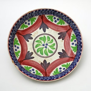 Pode incluir: Um prato redondo de cerâmica pintado à mão com base creme. O prato apresenta um desenho floral verde central, rodeado por formas de fita vermelha, detalhes roxos e uma borda azul estampada.